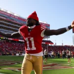 FootballR - NFL - Ein Footballspieler in einer rot-goldenen 49ers-Uniform mit der Nummer 11 streckt seine Arme in die Höhe, während er in einem großen Stadion über das Spielfeld läuft. Er trägt eine rote Kappe, hat einen Bart und sieht aus wie Brandon Aiyuk. Das Stadion ist voller Zuschauer, verschiedene Teambanner sind zu sehen und der Himmel ist klar und sonnig. Diese Beschreibung wurde automatisch generiert.