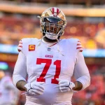 FootballR - NFL - Ein Footballspieler in weiß-roter Uniform, der Trent Williams ähnelt, steht auf dem Feld und trägt einen goldenen Helm mit getöntem Visier. Auf seinem Trikot ist die Nummer 71 mit Kapitänsaufnähern zu sehen, während er seine Handschuhe zurechtrückt. Im Hintergrund ist ein verschwommenes Stadion mit bunten Sitzplätzen zu sehen. Diese Beschreibung wurde automatisch generiert.