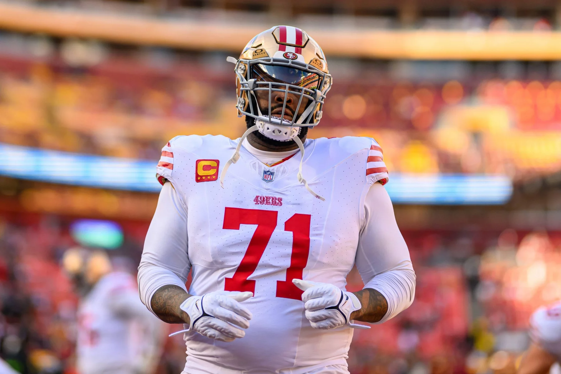 FootballR - NFL - Ein Footballspieler in weiß-roter Uniform, der Trent Williams ähnelt, steht auf dem Feld und trägt einen goldenen Helm mit getöntem Visier. Auf seinem Trikot ist die Nummer 71 mit Kapitänsaufnähern zu sehen, während er seine Handschuhe zurechtrückt. Im Hintergrund ist ein verschwommenes Stadion mit bunten Sitzplätzen zu sehen. Diese Beschreibung wurde automatisch generiert.