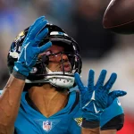 FootballR - NFL - Zay Jones, in einem blaugrünen Trikot und einem schwarzen Helm mit Jaguar-Logo sowie blauen Handschuhen, bereitet sich darauf vor, einen Football zu fangen. Seine Augen sind auf den Ball gerichtet und seine Hände sind nach oben ausgestreckt, um ihn zu fangen. Der unscharfe Hintergrund lässt auf ein überfülltes Stadion schließen. Diese Beschreibung wurde automatisch generiert.