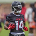 FootballR - NFL Rondale Moore - Ein Footballspieler mit schwarzem Falcons-Helm, schwarzem Trikot mit der Aufschrift „ATL“ in Rot und Handschuhen hält einen Football. Der Spieler steht während des Trainings auf einem Feld, andere Spieler und Trainer sind im Hintergrund verschwommen. Die Umgebung lässt auf Training oder eine Übungseinheit schließen. Diese Beschreibung wurde automatisch generiert.