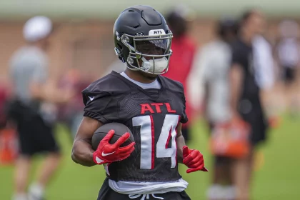 FootballR - NFL Rondale Moore - Ein Footballspieler mit schwarzem Falcons-Helm, schwarzem Trikot mit der Aufschrift „ATL“ in Rot und Handschuhen hält einen Football. Der Spieler steht während des Trainings auf einem Feld, andere Spieler und Trainer sind im Hintergrund verschwommen. Die Umgebung lässt auf Training oder eine Übungseinheit schließen. Diese Beschreibung wurde automatisch generiert.