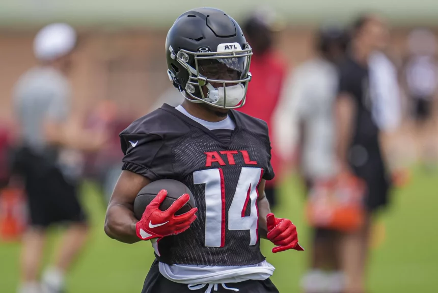 FootballR - NFL Rondale Moore - Ein Footballspieler mit schwarzem Falcons-Helm, schwarzem Trikot mit der Aufschrift „ATL“ in Rot und Handschuhen hält einen Football. Der Spieler steht während des Trainings auf einem Feld, andere Spieler und Trainer sind im Hintergrund verschwommen. Die Umgebung lässt auf Training oder eine Übungseinheit schließen. Diese Beschreibung wurde automatisch generiert.