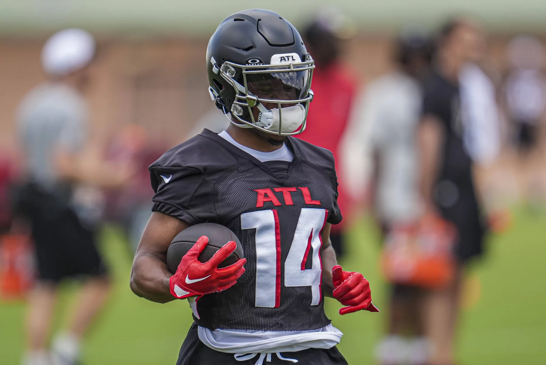 FootballR - NFL Rondale Moore - Ein Footballspieler mit schwarzem Falcons-Helm, schwarzem Trikot mit der Aufschrift „ATL“ in Rot und Handschuhen hält einen Football. Der Spieler steht während des Trainings auf einem Feld, andere Spieler und Trainer sind im Hintergrund verschwommen. Die Umgebung lässt auf Training oder eine Übungseinheit schließen. Diese Beschreibung wurde automatisch generiert.