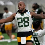 FootballR - NFL - Ein Footballspieler in einem weißen Packers-Trikot mit der Nummer 28 und grünen Shorts streckt in einer Trainingsanlage seine Arme aus. Hinter ihm kniet ein anderer Spieler in einem ähnlichen Outfit mit der Nummer 8 auf einem Knie. Im unscharfen Hintergrund der Trainingseinheit der NFL Saison 2024 mit AJ Dillon sind weitere Spieler und Ausrüstung zu sehen. Diese Beschreibung wurde automatisch generiert.