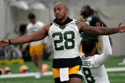 FootballR - NFL - Ein Footballspieler in einem weißen Packers-Trikot mit der Nummer 28 und grünen Shorts streckt in einer Trainingsanlage seine Arme aus. Hinter ihm kniet ein anderer Spieler in einem ähnlichen Outfit mit der Nummer 8 auf einem Knie. Im unscharfen Hintergrund der Trainingseinheit der NFL Saison 2024 mit AJ Dillon sind weitere Spieler und Ausrüstung zu sehen. Diese Beschreibung wurde automatisch generiert.