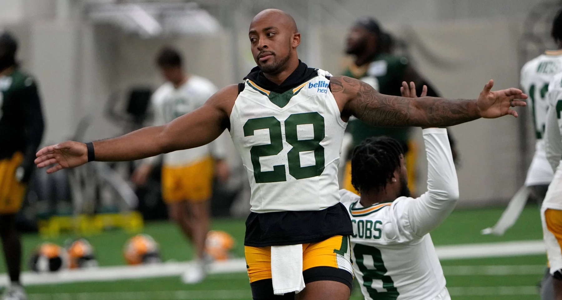 FootballR - NFL - Ein Footballspieler in einem weißen Packers-Trikot mit der Nummer 28 und grünen Shorts streckt in einer Trainingsanlage seine Arme aus. Hinter ihm kniet ein anderer Spieler in einem ähnlichen Outfit mit der Nummer 8 auf einem Knie. Im unscharfen Hintergrund der Trainingseinheit der NFL Saison 2024 mit AJ Dillon sind weitere Spieler und Ausrüstung zu sehen. Diese Beschreibung wurde automatisch generiert.