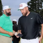 FootballR - NFL New Heights - Zwei Männer mit Baseballkappen stehen draußen. Der Mann links trägt ein grünes Poloshirt und beige Hosen, eine weiße Kappe mit grünem Logo und einen Vollbart. Der Mann rechts trägt ein schwarzes Nike-Poloshirt und weiße Hosen, hält ein Telefon in der Hand und trägt stolz seine Kelce-Brüder-Kappe mit rotem Logo. Diese Beschreibung wurde automatisch generiert.