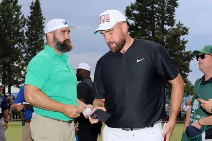 FootballR - NFL New Heights - Zwei Männer mit Baseballkappen stehen draußen. Der Mann links trägt ein grünes Poloshirt und beige Hosen, eine weiße Kappe mit grünem Logo und einen Vollbart. Der Mann rechts trägt ein schwarzes Nike-Poloshirt und weiße Hosen, hält ein Telefon in der Hand und trägt stolz seine Kelce-Brüder-Kappe mit rotem Logo. Diese Beschreibung wurde automatisch generiert.