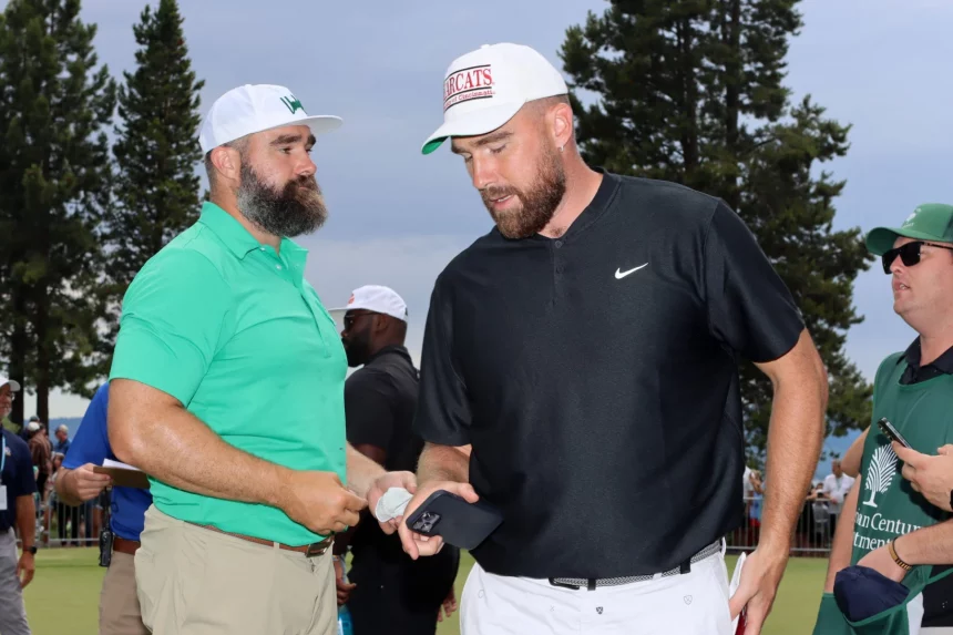 FootballR - NFL New Heights - Zwei Männer mit Baseballkappen stehen draußen. Der Mann links trägt ein grünes Poloshirt und beige Hosen, eine weiße Kappe mit grünem Logo und einen Vollbart. Der Mann rechts trägt ein schwarzes Nike-Poloshirt und weiße Hosen, hält ein Telefon in der Hand und trägt stolz seine Kelce-Brüder-Kappe mit rotem Logo. Diese Beschreibung wurde automatisch generiert.