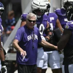 FootballR - NFL - OL-Trainer Joe D'Alessandris, in einem lila Ravens-Shirt und Sonnenbrille, steht mit ausgestreckter rechter Hand zwischen Footballspielern. Die Spieler tragen lila Trikots, weiße Hosen und Helme. Der Spieler rechts, Nummer 79, unterhält sich mit Joe. Im Hintergrund sind andere Spieler und Personen zu sehen. Diese Beschreibung wurde automatisch generiert.