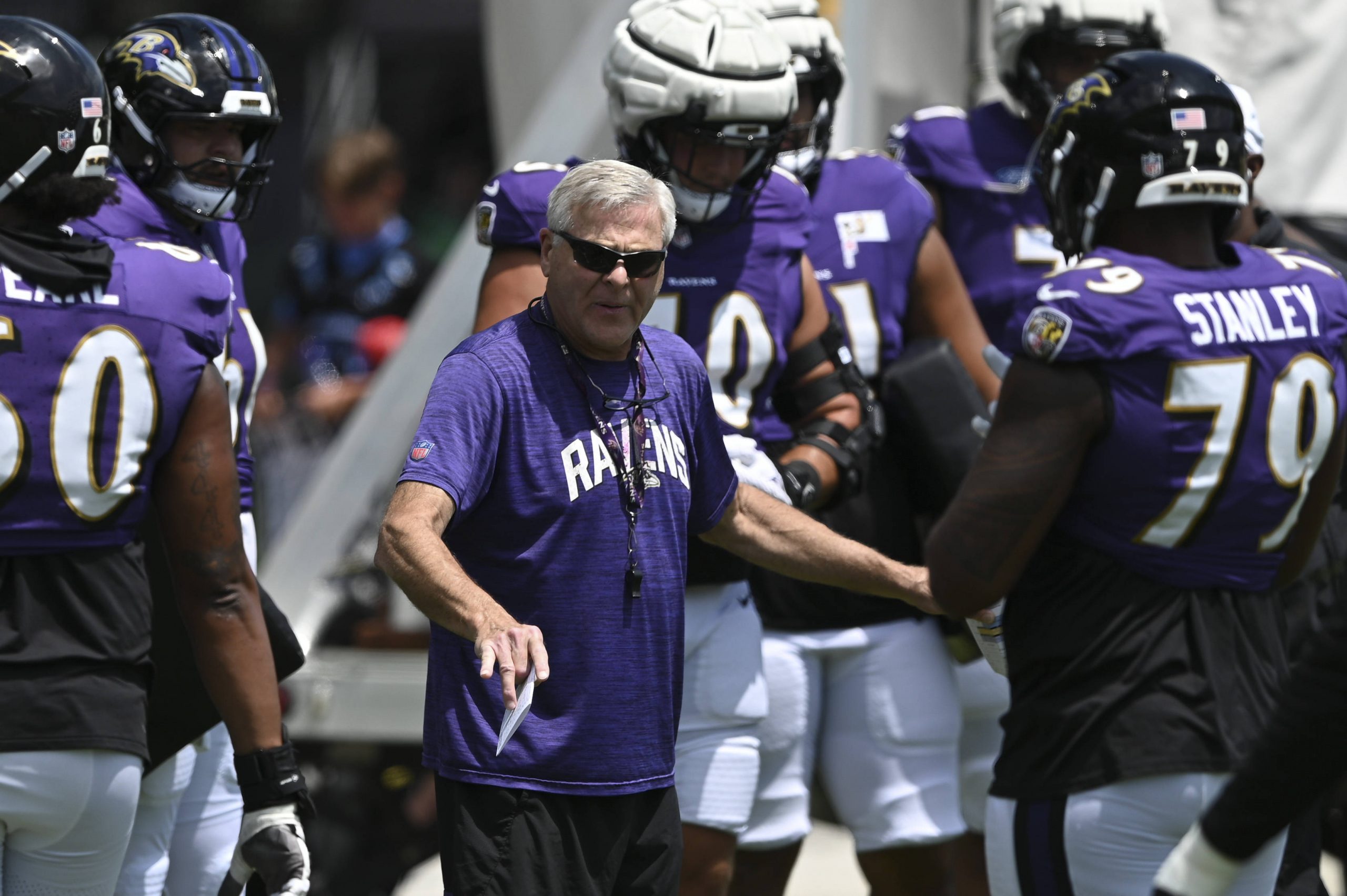 FootballR - NFL - OL-Trainer Joe D'Alessandris, in einem lila Ravens-Shirt und Sonnenbrille, steht mit ausgestreckter rechter Hand zwischen Footballspielern. Die Spieler tragen lila Trikots, weiße Hosen und Helme. Der Spieler rechts, Nummer 79, unterhält sich mit Joe. Im Hintergrund sind andere Spieler und Personen zu sehen. Diese Beschreibung wurde automatisch generiert.