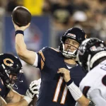 FootballR - NFL - Ein Football-Quarterback in einer marineblauen Bears-Uniform mit der Nummer 11 auf seinem Trikot ist mitten im Wurf und hält den Ball während des Hall of Fame Game 2024 hoch in seiner rechten Hand. Ein Offensive Lineman in einer ähnlichen Uniform blockt einen Defensivspieler in einem weißen Trikot mit der Nummer 99, der auf den Quarterback zustürmt. Im Hintergrund ist eine Menge zu sehen, die aufsteht und zusieht Diese Beschreibung wurde automatisch generiert.