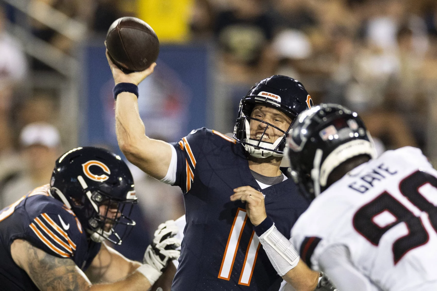 FootballR - NFL - Ein Football-Quarterback in einer marineblauen Bears-Uniform mit der Nummer 11 auf seinem Trikot ist mitten im Wurf und hält den Ball während des Hall of Fame Game 2024 hoch in seiner rechten Hand. Ein Offensive Lineman in einer ähnlichen Uniform blockt einen Defensivspieler in einem weißen Trikot mit der Nummer 99, der auf den Quarterback zustürmt. Im Hintergrund ist eine Menge zu sehen, die aufsteht und zusieht Diese Beschreibung wurde automatisch generiert.