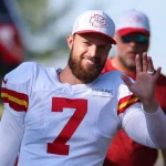FootballR - NFL - Ein Footballspieler, möglicherweise Chiefs-Kicker Harrison Butker, trägt ein weißes Trikot mit der Nummer 7 und eine passende Kappe und winkt lächelnd. Er hat einen Bart und einen sichtbaren Ehering. Eine weitere Person im Hintergrund trägt ein rotes Hemd und eine Sonnenbrille. Die Szene scheint im Freien mit einem verschwommenen grünen Hintergrund zu sein. Diese Beschreibung wurde automatisch generiert.
