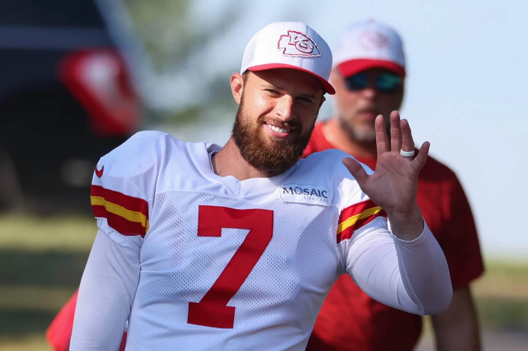 FootballR - NFL - Ein Footballspieler, möglicherweise Chiefs-Kicker Harrison Butker, trägt ein weißes Trikot mit der Nummer 7 und eine passende Kappe und winkt lächelnd. Er hat einen Bart und einen sichtbaren Ehering. Eine weitere Person im Hintergrund trägt ein rotes Hemd und eine Sonnenbrille. Die Szene scheint im Freien mit einem verschwommenen grünen Hintergrund zu sein. Diese Beschreibung wurde automatisch generiert.