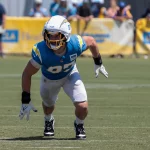 FootballR - NFL - Ein Footballspieler, der Joey Bosa ähnelt, ein blaues Trikot mit der Nummer 97 und einen weißen Helm trägt, steht in athletischer Haltung auf einem Feld. Er scheint sich mitten in einer Bewegung zu befinden und den Blick nach vorne gerichtet zu halten. Im Hintergrund sind andere Spieler, Trainer und ein gelbes Banner mit der Aufschrift „UCLA“ zu sehen. Es ist eine sonnige Trainingseinheit im Freien. Diese Beschreibung wurde automatisch generiert.