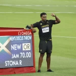 FootballR - NFL - Ein männlicher Athlet steht neben einer elektronischen Anzeigetafel mit der Aufschrift „Männer-Diskuswurf“, wobei „NEW OR“ und „JAM 70.00 STONA R“ einen neuen olympischen Rekord anzeigen. Diskus-Champion Roje Stona, der ein schwarzes Outfit mit „JAMAICA“ und „STONA“ auf seiner Startnummer trägt, spannt beim Grasfeld-Event seinen rechten Bizeps an Diese Beschreibung wurde automatisch generiert.