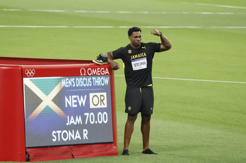 FootballR - NFL - Ein männlicher Athlet steht neben einer elektronischen Anzeigetafel mit der Aufschrift „Männer-Diskuswurf“, wobei „NEW OR“ und „JAM 70.00 STONA R“ einen neuen olympischen Rekord anzeigen. Diskus-Champion Roje Stona, der ein schwarzes Outfit mit „JAMAICA“ und „STONA“ auf seiner Startnummer trägt, spannt beim Grasfeld-Event seinen rechten Bizeps an Diese Beschreibung wurde automatisch generiert.