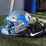 FootballR - NFL - Ein silber-blauer Footballhelm der Detroit Lions liegt auf einem Rasen neben einem Paar schwarzer Nike-Footballschuhe und Handschuhe. Der Helm, auf dem der stilisierte blaue Löwe im Sprung abgebildet ist, liegt zwischen Kabeln und Ausrüstung im verschwommenen Hintergrund. Eine Erinnerung daran, wie der deutsche NFL-Profi David Bada einst einer schweren Verletzung entging. Diese Beschreibung wurde automatisch generiert.