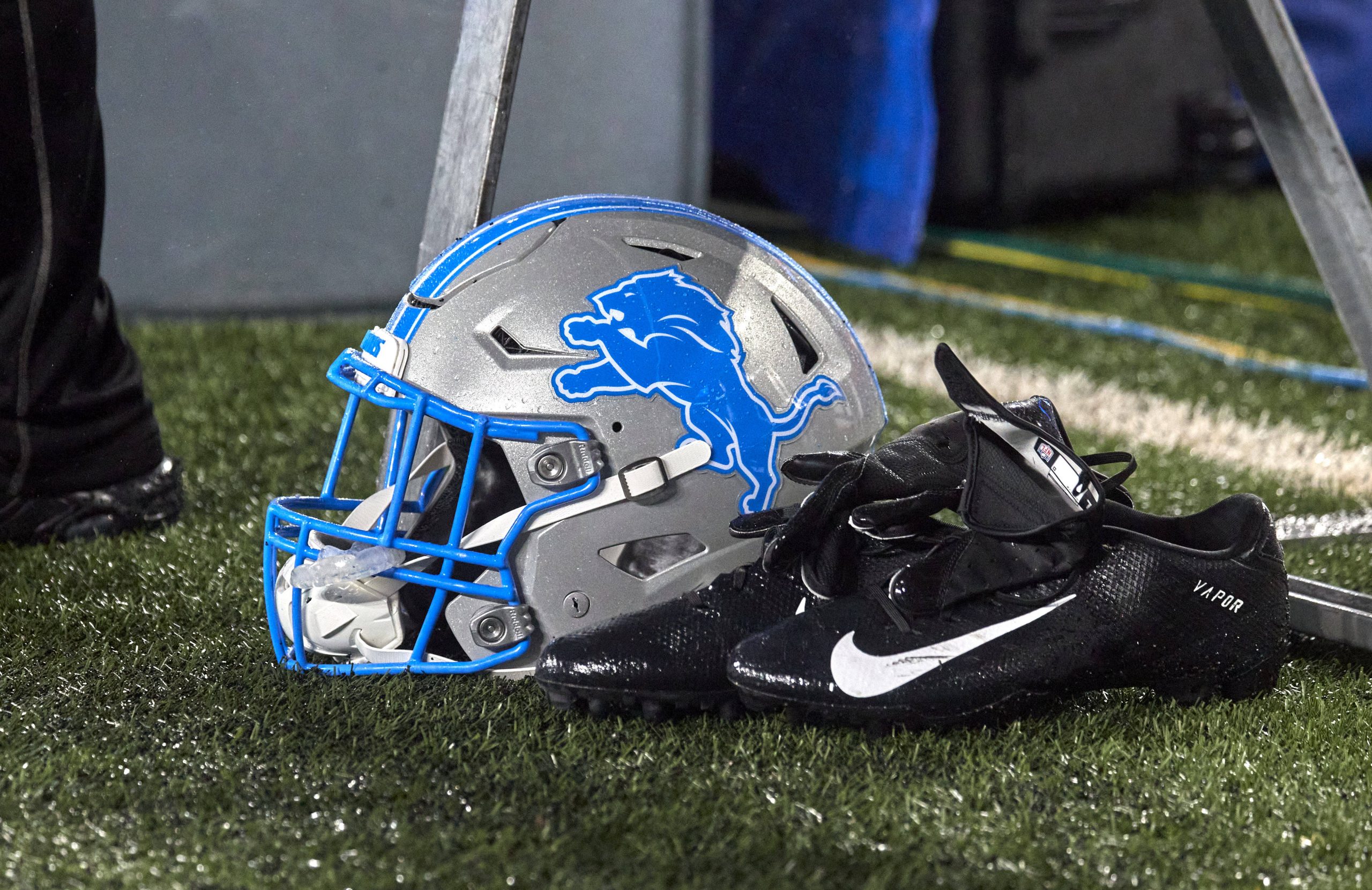 FootballR - NFL - Ein silber-blauer Footballhelm der Detroit Lions liegt auf einem Rasen neben einem Paar schwarzer Nike-Footballschuhe und Handschuhe. Der Helm, auf dem der stilisierte blaue Löwe im Sprung abgebildet ist, liegt zwischen Kabeln und Ausrüstung im verschwommenen Hintergrund. Eine Erinnerung daran, wie der deutsche NFL-Profi David Bada einst einer schweren Verletzung entging. Diese Beschreibung wurde automatisch generiert.