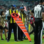 FootballR - NFL Hawk Eye - Zwei Schiedsrichter an der Seitenlinie, die gelbe Westen und Mützen tragen, halten während eines Footballspiels NFL-Markierungen fest. Der Schiedsrichter im Vordergrund hält eine leuchtend orange Markierung mit den Zahlen „2“ und „3“. In der Nähe steht ein Schiedsrichter in einem schwarz-weiß gestreiften Hemd. Im Hintergrund sind Cheerleader und Spieler zu sehen, die die Hightech-Revolution der NFL hervorheben. Diese Beschreibung wurde automatisch generiert.