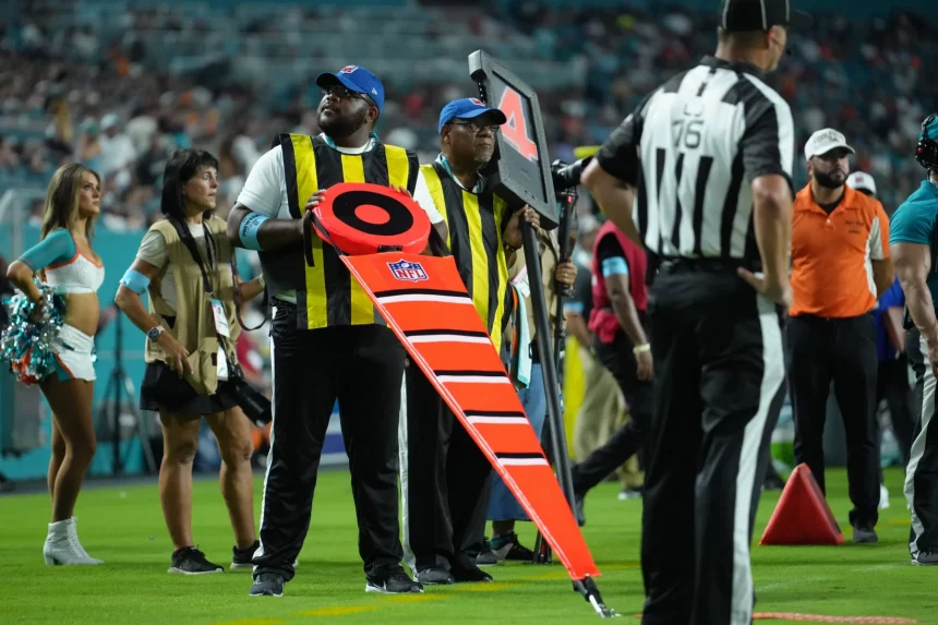 FootballR - NFL Hawk Eye - Zwei Schiedsrichter an der Seitenlinie, die gelbe Westen und Mützen tragen, halten während eines Footballspiels NFL-Markierungen fest. Der Schiedsrichter im Vordergrund hält eine leuchtend orange Markierung mit den Zahlen „2“ und „3“. In der Nähe steht ein Schiedsrichter in einem schwarz-weiß gestreiften Hemd. Im Hintergrund sind Cheerleader und Spieler zu sehen, die die Hightech-Revolution der NFL hervorheben. Diese Beschreibung wurde automatisch generiert.