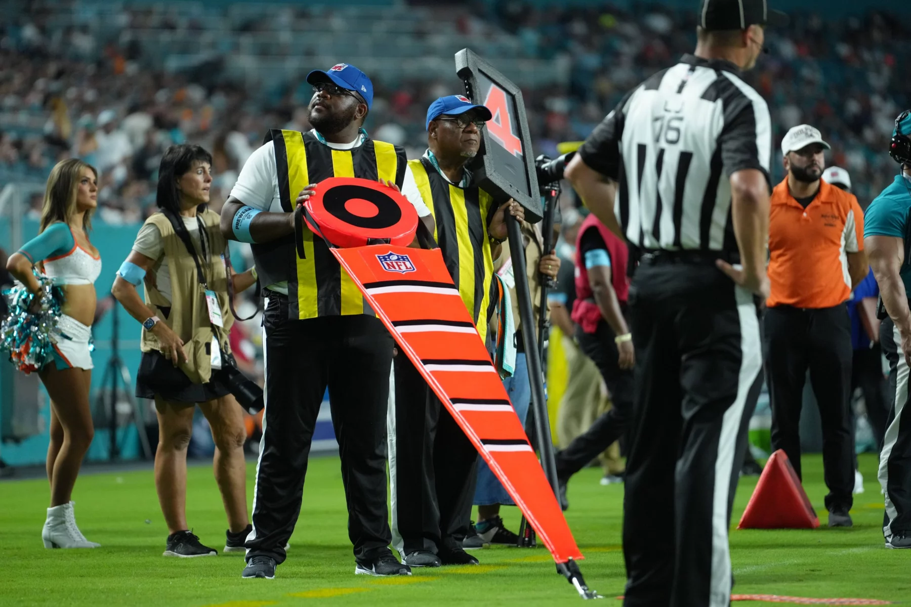 FootballR - NFL Hawk Eye - Zwei Schiedsrichter an der Seitenlinie, die gelbe Westen und Mützen tragen, halten während eines Footballspiels NFL-Markierungen fest. Der Schiedsrichter im Vordergrund hält eine leuchtend orange Markierung mit den Zahlen „2“ und „3“. In der Nähe steht ein Schiedsrichter in einem schwarz-weiß gestreiften Hemd. Im Hintergrund sind Cheerleader und Spieler zu sehen, die die Hightech-Revolution der NFL hervorheben. Diese Beschreibung wurde automatisch generiert.