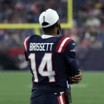 FootballR - NFL Drake Maye oder Jacoby Brissett - Ein Footballspieler steht auf dem Spielfeld. Er trägt ein marineblaues Patriots-Trikot mit roten und weißen Streifen und dem Namen Jacoby Brissett sowie der Nummer 14 auf dem Rücken. Er trägt eine weiße Kappe verkehrt herum und scheint das Spiel mit verschränkten Armen zu beobachten. Das Stadion ist voller Zuschauer. Diese Beschreibung wurde automatisch generiert.