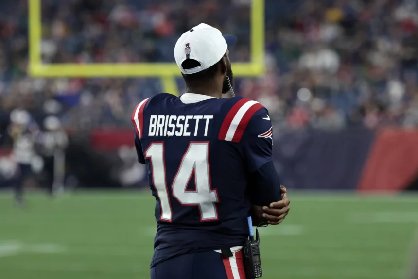 FootballR - NFL Drake Maye oder Jacoby Brissett - Ein Footballspieler steht auf dem Spielfeld. Er trägt ein marineblaues Patriots-Trikot mit roten und weißen Streifen und dem Namen Jacoby Brissett sowie der Nummer 14 auf dem Rücken. Er trägt eine weiße Kappe verkehrt herum und scheint das Spiel mit verschränkten Armen zu beobachten. Das Stadion ist voller Zuschauer. Diese Beschreibung wurde automatisch generiert.