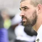 FootballR - NFL - Ein Mann mit kurzen Haaren und Bart, der ein weißes Hemd mit dem Ravens-Star-Logo trägt, blickt in die Ferne. Der Hintergrund ist verschwommen, was seinen nachdenklichen Gesichtsausdruck betont. Die Umgebung lässt vermuten, dass er sich auf einer Sportveranstaltung befindet und möglicherweise über die letzten Spiele oder Ereignisse mit Mark Andrews nachdenkt. Diese Beschreibung wurde automatisch generiert.
