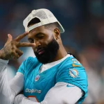 FootballR - NFL - Ein Footballspieler in der Uniform der Miami Dolphins posiert mit dem linken Arm vor der Brust und der rechten Hand, die über seinem rechten Auge ein Peace-Zeichen macht. Er trägt einen Bart und eine weiße, verkehrt herum aufgesetzte Kappe und strahlt ein Selbstvertrauen aus, das an Odell Beckham Jr. erinnert. Im Hintergrund ist ein unscharfes Stadion zu sehen. Diese Beschreibung wurde automatisch generiert.