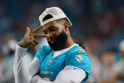 FootballR - NFL - Ein Footballspieler in der Uniform der Miami Dolphins posiert mit dem linken Arm vor der Brust und der rechten Hand, die über seinem rechten Auge ein Peace-Zeichen macht. Er trägt einen Bart und eine weiße, verkehrt herum aufgesetzte Kappe und strahlt ein Selbstvertrauen aus, das an Odell Beckham Jr. erinnert. Im Hintergrund ist ein unscharfes Stadion zu sehen. Diese Beschreibung wurde automatisch generiert.