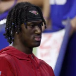 FootballR - NFL Marvin Harrison Jr. vs Fanatics - Eine Person mit dunklen geflochtenen Haaren, einer roten Kapuzenjacke und einem schwarzen Stirnband mit einem roten Vogellogo ist zu sehen. Sie blickt mit neutralem Gesichtsausdruck nach vorne. Der Hintergrund ist verschwommen und zeigt Menschen und undeutliche Beschilderungen – vielleicht ist es ein Fan der NFL-Legende Harrison oder ein Rookie-Sohn. Diese Beschreibung wurde automatisch generiert.