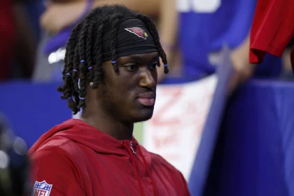 FootballR - NFL Marvin Harrison Jr. vs Fanatics - Eine Person mit dunklen geflochtenen Haaren, einer roten Kapuzenjacke und einem schwarzen Stirnband mit einem roten Vogellogo ist zu sehen. Sie blickt mit neutralem Gesichtsausdruck nach vorne. Der Hintergrund ist verschwommen und zeigt Menschen und undeutliche Beschilderungen – vielleicht ist es ein Fan der NFL-Legende Harrison oder ein Rookie-Sohn. Diese Beschreibung wurde automatisch generiert.