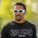 FootballR - NFL - Ein Mann mit lockigem dunklem Haar und Spitzbart trägt weiße, umlaufende Sonnenbrillen und ein schwarzes T-Shirt mit dem Text „Jakob Johnson“ in stilisierter Schrift. Er steht im Freien mit verschwommenem Grün im Hintergrund. Trotz Deutscher Hoffnungen deutet sein entschlossener Blick auf drohende NFL Roster Cuts hin. Außerdem trägt er einen kleinen Ohrring und eine schwarze Halskette. Deutsche Hoffnungen zerschlagen. Diese Beschreibung wurde automatisch generiert.
