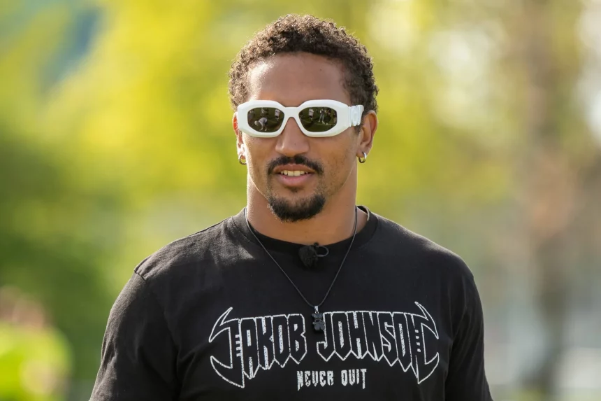 FootballR - NFL - Ein Mann mit lockigem dunklem Haar und Spitzbart trägt weiße, umlaufende Sonnenbrillen und ein schwarzes T-Shirt mit dem Text „Jakob Johnson“ in stilisierter Schrift. Er steht im Freien mit verschwommenem Grün im Hintergrund. Trotz Deutscher Hoffnungen deutet sein entschlossener Blick auf drohende NFL Roster Cuts hin. Außerdem trägt er einen kleinen Ohrring und eine schwarze Halskette. Deutsche Hoffnungen zerschlagen. Diese Beschreibung wurde automatisch generiert.