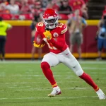 FootballR - NFL - Ein Footballspieler der Kansas City Chiefs in rot-weißer Uniform und mit Helm läuft während eines Spiels mit dem Ball. Kadarius Toney hält den Ball in beiden Händen und trägt gelbe Handschuhe und Stollenschuhe. Im Hintergrund sind verschwommene Zuschauer und Mannschaftsmitglieder am Spielfeldrand zu sehen. Diese Beschreibung wurde automatisch generiert.