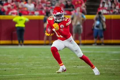 FootballR - NFL - Ein Footballspieler der Kansas City Chiefs in rot-weißer Uniform und mit Helm läuft während eines Spiels mit dem Ball. Kadarius Toney hält den Ball in beiden Händen und trägt gelbe Handschuhe und Stollenschuhe. Im Hintergrund sind verschwommene Zuschauer und Mannschaftsmitglieder am Spielfeldrand zu sehen. Diese Beschreibung wurde automatisch generiert.