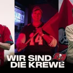 FootballR - NFL - Drei Personen stehen in einer Reihe und halten Buccaneers-Fahnen. Sie tragen unterschiedliche Buccaneers-Kleidung: ein Trikot mit der Aufschrift „55“, ein Shirt mit der Aufschrift „14“ und einen hellen Rundhalsausschnitt. Im Hintergrund sind ein Bildschirm und ein Sonnenuntergang zu sehen. Unten sind „Wir Sind Die Krewe“ und das Logo der Tampa Bay Buccaneers zu sehen, was das Engagement in Deutschland zeigt. Diese Beschreibung wurde automatisch generiert.