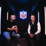 FootballR - NFL - Zwei Männer sitzen in einem schwach beleuchteten Raum mit blauen und roten vertikalen Lichtern auf beiden Seiten. Ein Mann auf der linken Seite hält einen Football und trägt ein schwarzes Outfit. Der Mann auf der rechten Seite, gekleidet in ein weißes Hemd und eine dunkle Weste, lächelt. Hinter ihnen hebt ein leuchtendes Neon-NFL-Logo Bitpanda als offiziellen Partner hervor. Diese Beschreibung wurde automatisch generiert.