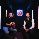 FootballR - NFL - Zwei Männer sitzen in einem schwach beleuchteten Raum mit blauen und roten vertikalen Lichtern auf beiden Seiten. Ein Mann auf der linken Seite hält einen Football und trägt ein schwarzes Outfit. Der Mann auf der rechten Seite, gekleidet in ein weißes Hemd und eine dunkle Weste, lächelt. Hinter ihnen hebt ein leuchtendes Neon-NFL-Logo Bitpanda als offiziellen Partner hervor. Diese Beschreibung wurde automatisch generiert.