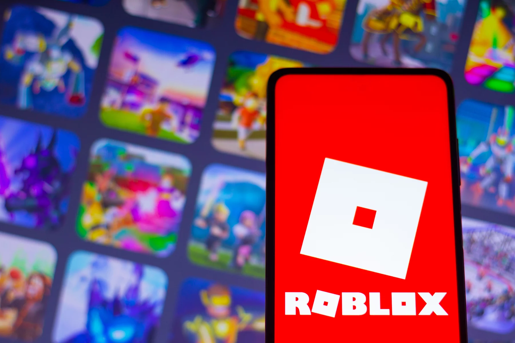 FootballR - NFL - Ein Smartphone mit dem Roblox-Logo, einem weißen, schrägen Quadrat mit einem kleineren, in der Mitte ausgeschnittenen Quadrat auf einem leuchtend roten Hintergrund. Hinter dem Telefon ist der Hintergrund verschwommen, zeigt aber bunte Spielsymbole, darunter NFL Universe Football, die verschiedene Auswahlmöglichkeiten auf der Roblox-Plattform hervorheben. Diese Beschreibung wurde automatisch generiert.