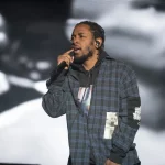 FootballR - NFL Kendrick Lamar - Ein Mann in einem karierten Hemd tritt auf der Bühne auf und hält dabei ein Mikrofon in der Hand. Er singt mitten im Gesang und hat einen angespannten Gesichtsausdruck. Im Hintergrund ist ein großes Schwarzweißbild eines anderen Gesichts zu sehen, das leicht unscharf ist und dem Bühnenbild während der Halbzeitshow des Super Bowl LIX Tiefe verleiht. Diese Beschreibung wurde automatisch generiert.