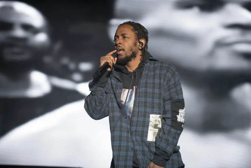 FootballR - NFL Kendrick Lamar - Ein Mann in einem karierten Hemd tritt auf der Bühne auf und hält dabei ein Mikrofon in der Hand. Er singt mitten im Gesang und hat einen angespannten Gesichtsausdruck. Im Hintergrund ist ein großes Schwarzweißbild eines anderen Gesichts zu sehen, das leicht unscharf ist und dem Bühnenbild während der Halbzeitshow des Super Bowl LIX Tiefe verleiht. Diese Beschreibung wurde automatisch generiert.