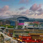 FootballR - NFL -Seattle Seahawks - Throwback Jerseys - Eine lebendige urbane Szene mit zwei nebeneinanderliegenden Stadien mit einzigartigen geschwungenen Dächern vor einem teilweise bewölkten Himmel. Darunter sind verschiedene Gebäude mit farbenfrohen Fassaden zu sehen, darunter ein markantes rotes Gebäude mit der Aufschrift „Miners Landing“. Im Vordergrund sind Straßen mit Bäumen und Fahrzeugen zu sehen, einige davon mit Retro-Trikots der Seattle Seahawks. Diese Beschreibung wurde automatisch generiert.