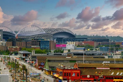 FootballR - NFL -Seattle Seahawks - Throwback Jerseys - Eine lebendige urbane Szene mit zwei nebeneinanderliegenden Stadien mit einzigartigen geschwungenen Dächern vor einem teilweise bewölkten Himmel. Darunter sind verschiedene Gebäude mit farbenfrohen Fassaden zu sehen, darunter ein markantes rotes Gebäude mit der Aufschrift „Miners Landing“. Im Vordergrund sind Straßen mit Bäumen und Fahrzeugen zu sehen, einige davon mit Retro-Trikots der Seattle Seahawks. Diese Beschreibung wurde automatisch generiert.