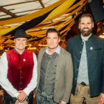 FootballR - NFL - Drei Männer in traditioneller bayerischer Kleidung stehen zusammen in einem für das Oktoberfest geschmückten Innenraum. Der Mann links trägt eine rote Weste und einen schwarzen Hut, der Mann in der Mitte einen braunen Blazer und ein weißes Hemd und der Mann rechts trägt eine grüne Jacke. Festliche Lichter und Banner schmücken den Hintergrund – wie ein automatisch gespeicherter Entwurf einer Feier. Diese Beschreibung wurde automatisch generiert.