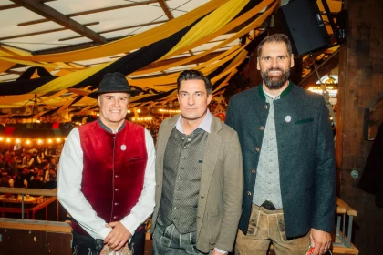 FootballR - NFL - Drei Männer in traditioneller bayerischer Kleidung stehen zusammen in einem für das Oktoberfest geschmückten Innenraum. Der Mann links trägt eine rote Weste und einen schwarzen Hut, der Mann in der Mitte einen braunen Blazer und ein weißes Hemd und der Mann rechts trägt eine grüne Jacke. Festliche Lichter und Banner schmücken den Hintergrund – wie ein automatisch gespeicherter Entwurf einer Feier. Diese Beschreibung wurde automatisch generiert.