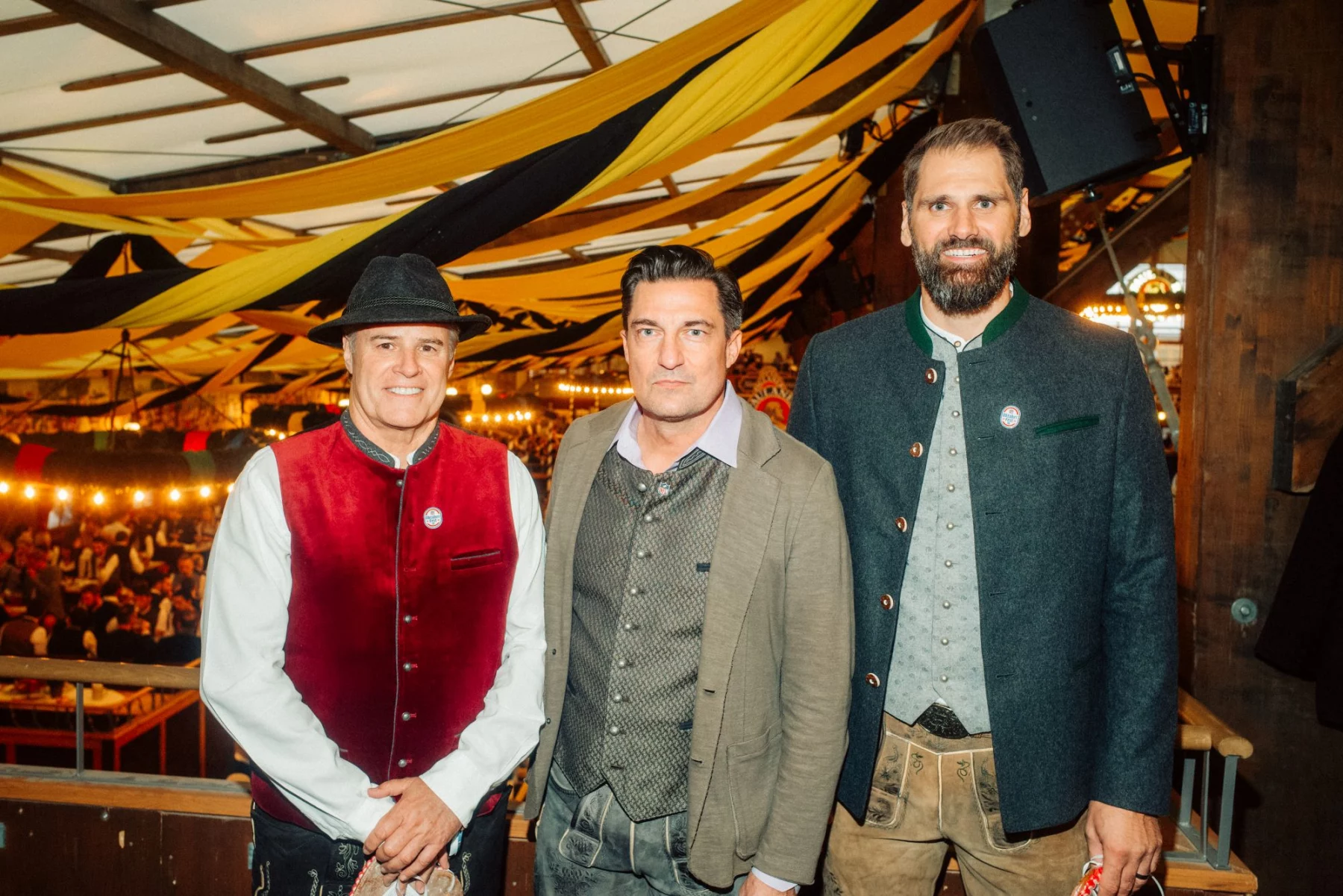FootballR - NFL - Drei Männer in traditioneller bayerischer Kleidung stehen zusammen in einem für das Oktoberfest geschmückten Innenraum. Der Mann links trägt eine rote Weste und einen schwarzen Hut, der Mann in der Mitte einen braunen Blazer und ein weißes Hemd und der Mann rechts trägt eine grüne Jacke. Festliche Lichter und Banner schmücken den Hintergrund – wie ein automatisch gespeicherter Entwurf einer Feier. Diese Beschreibung wurde automatisch generiert.
